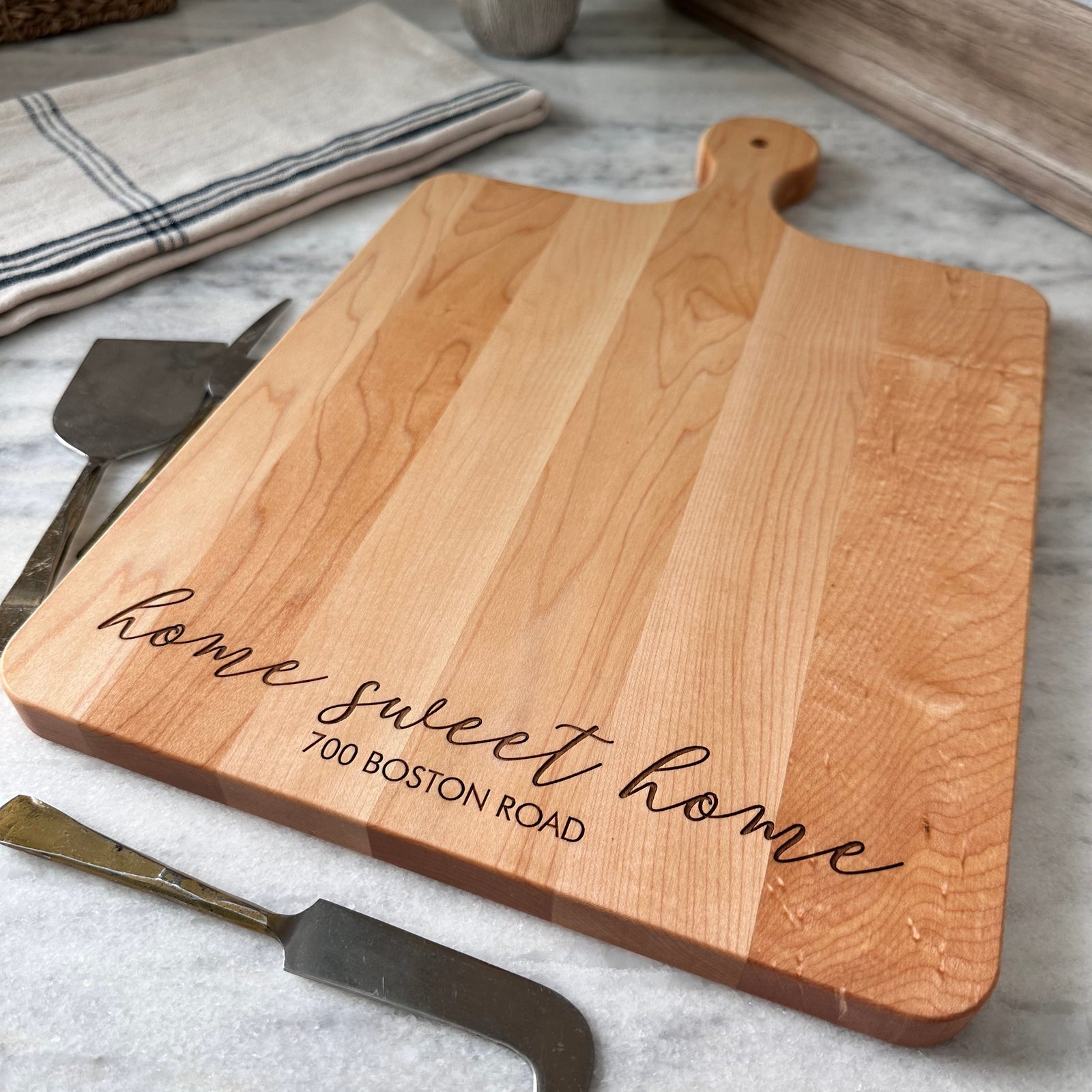 A personalized wooden charcuterie board with an engraved message, accompanied by a cheese knife and fork, presented on a kitchen counter.